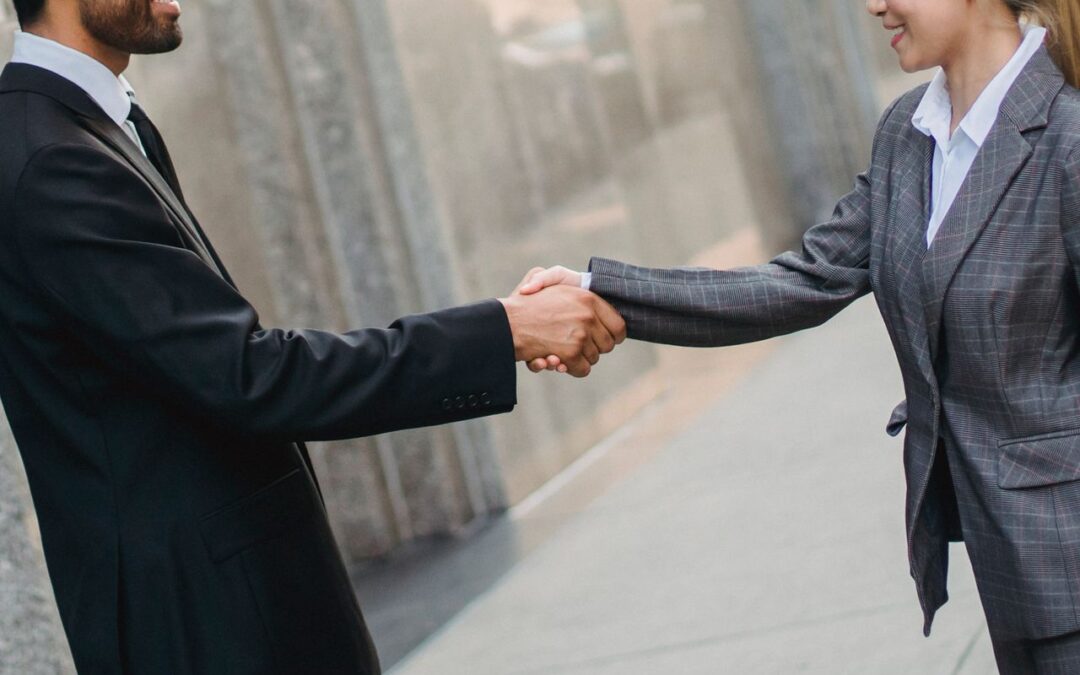 Two business professionals shaking hands outdoors, one in a black suit and tie, the other in a gray plaid blazer.